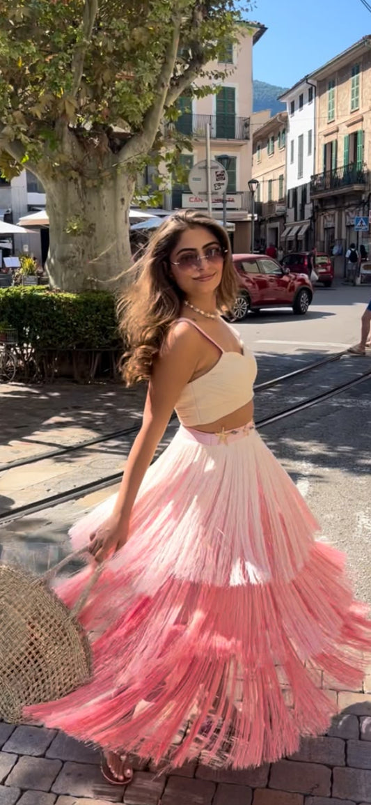 A woman wearing a two-piece pastel pink and cream crop top and pleated maxi skirt set, holding a straw hat, posing on a sunny outdoor street.
